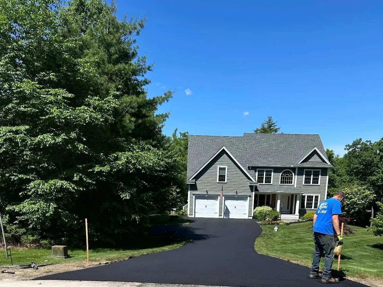 A worker in a blue shirt finishes spreading fresh, black asphalt for a residential driveway leading to a two-story home.