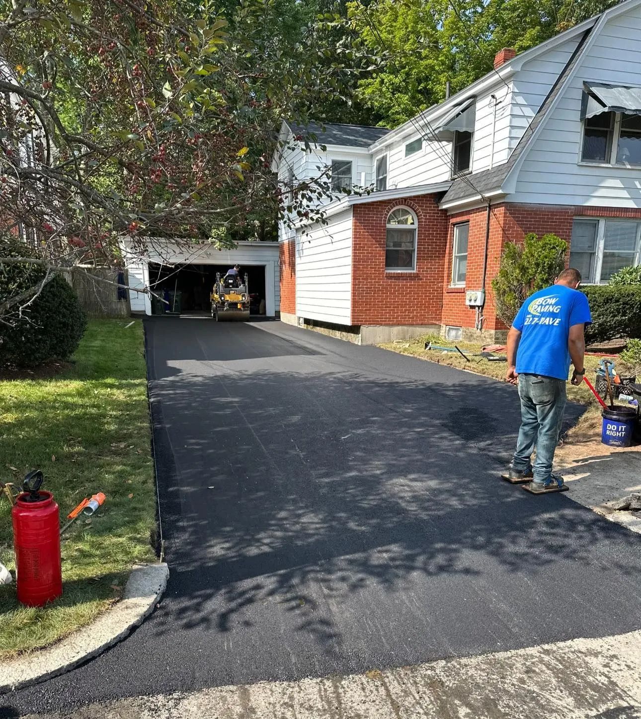 A worker in a blue shirt stands on a freshly paved black asphalt driveway leading to a garage next to a brick house.
