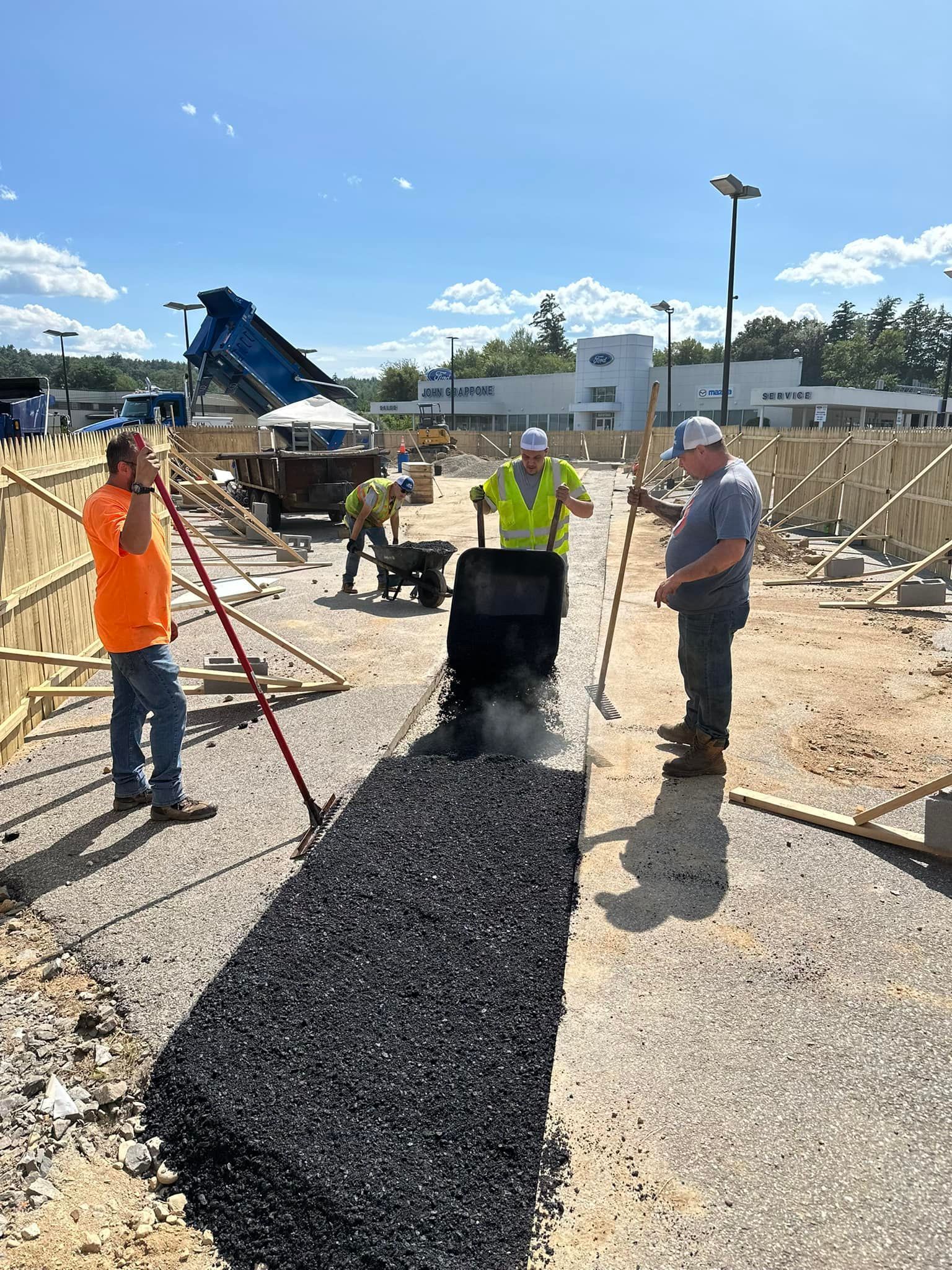Construction workers fill a trench with dark gravel at a sunny outdoor site with equipment in the background.