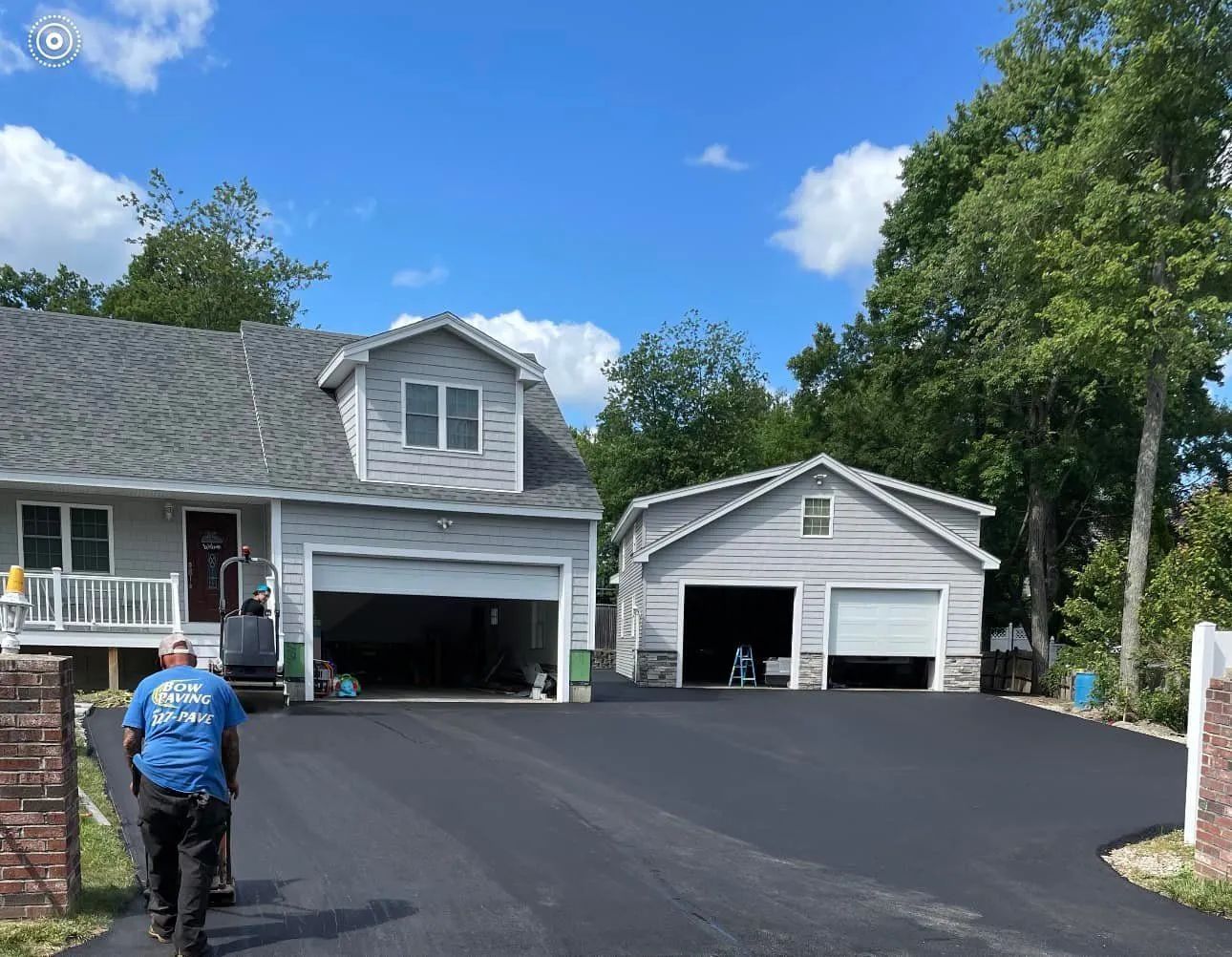 A worker uses a vibrating plate compactor to smooth a freshly paved asphalt driveway leading to a house and garage.