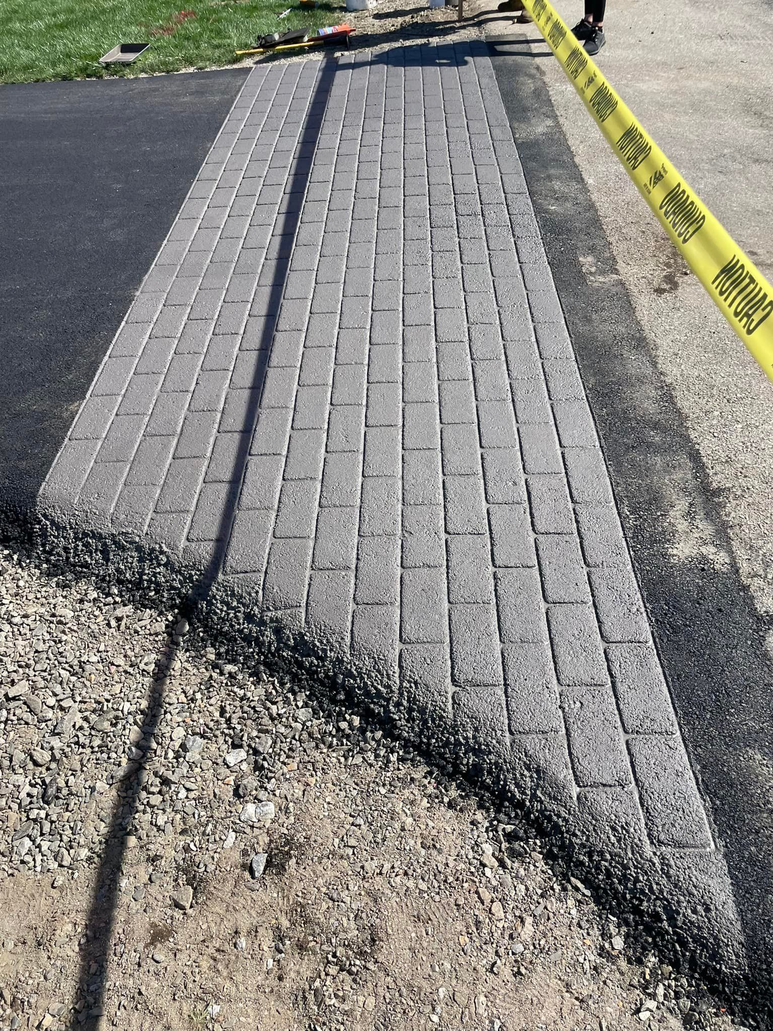 A rectangular section of gray pavers installed between asphalt paths, bordered by yellow caution tape.