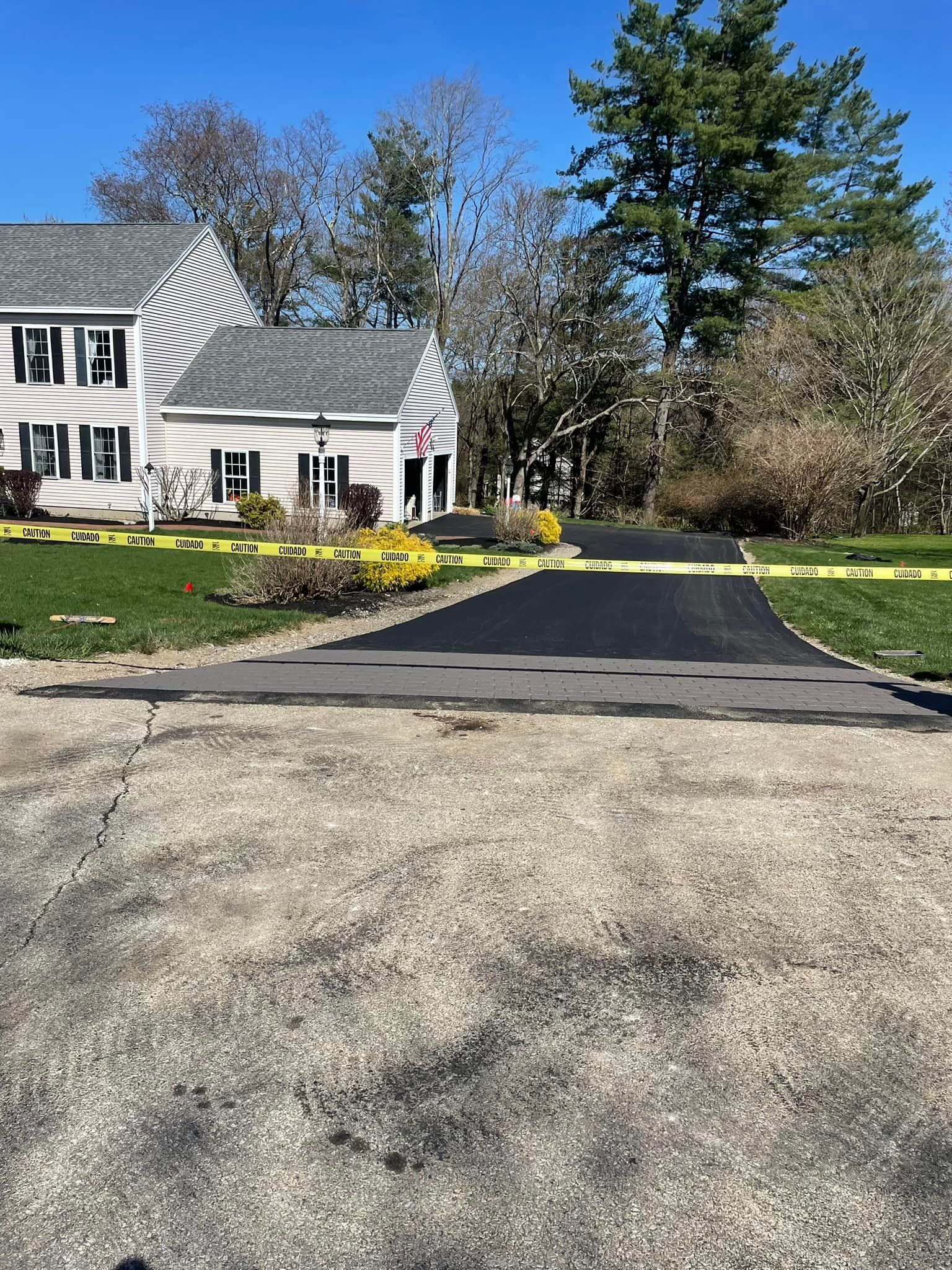 A paved driveway leading to a colonial-style house, bordered by yellow caution tape on a bright, sunny day.