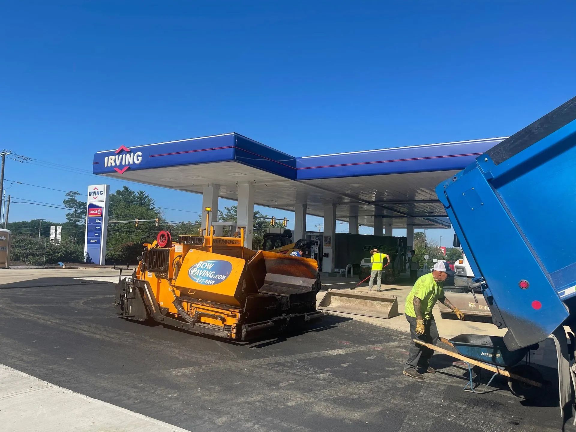 Construction workers use a yellow asphalt paver to pave a driveway at an Irving gas station on a sunny day.