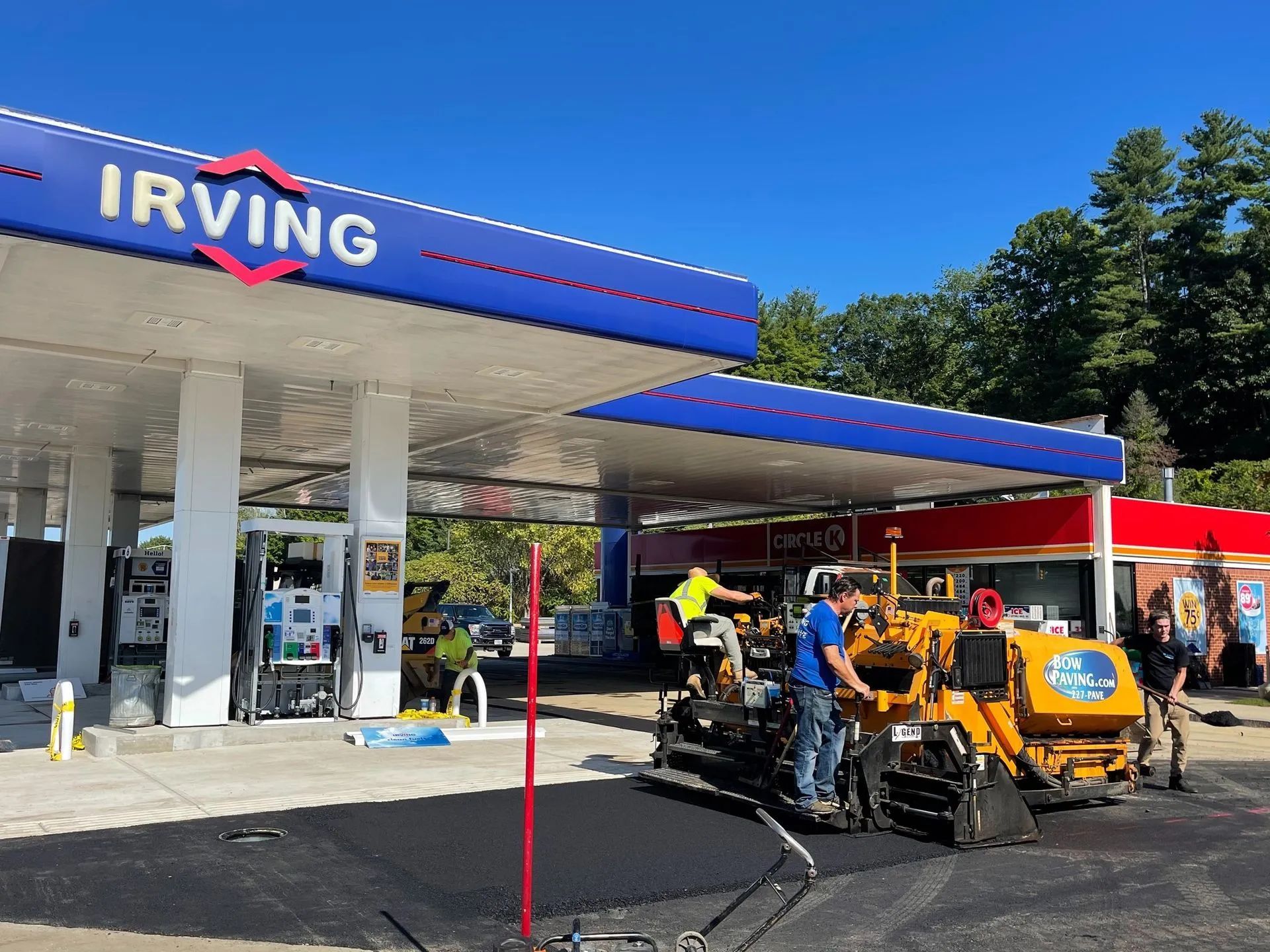 Construction workers operate yellow paving machinery to resurface the ground at an Irving gas station on a sunny day.