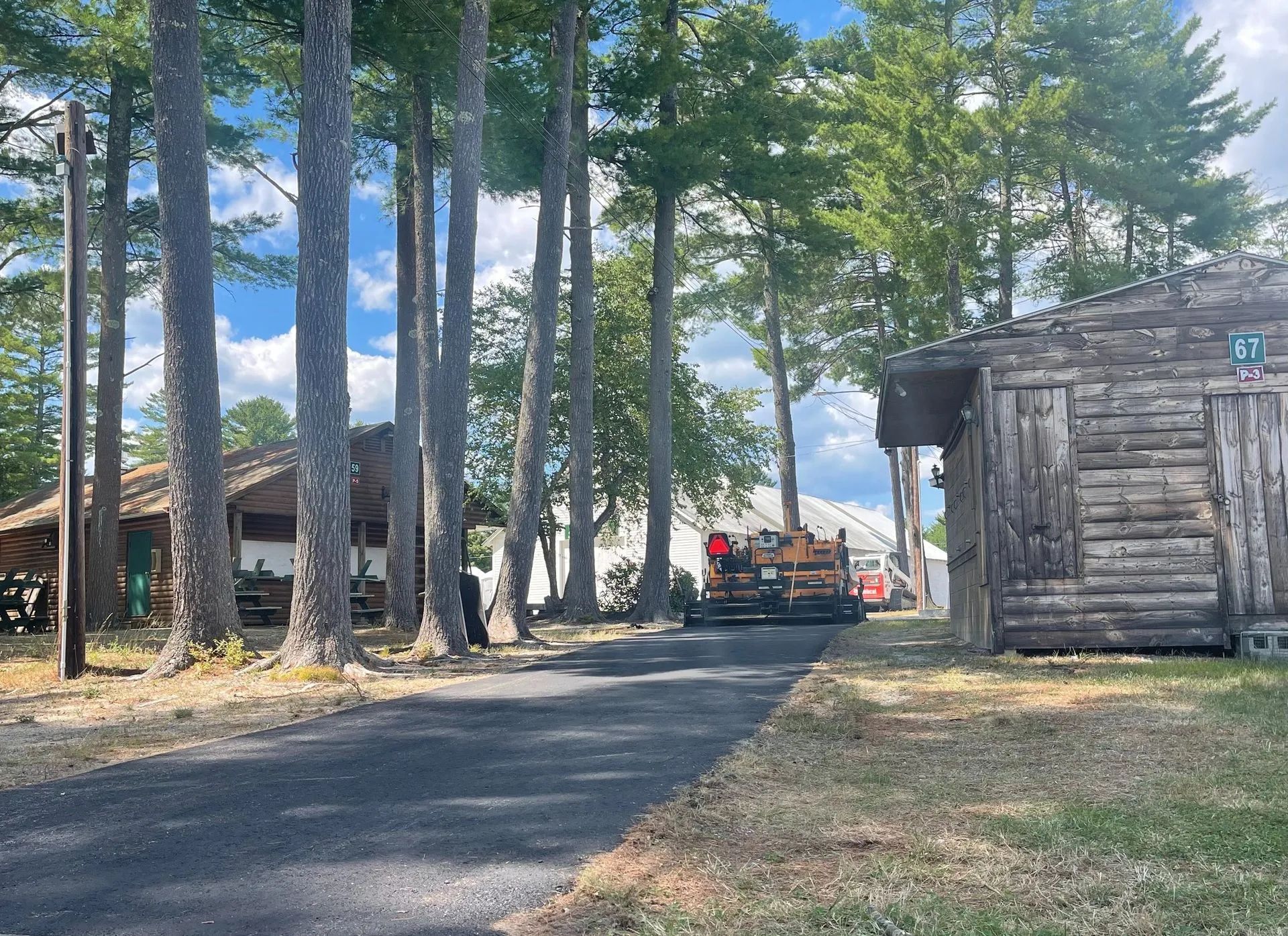 A paved path leads between tall pine trees toward a construction vehicle, with rustic log cabins on either side.