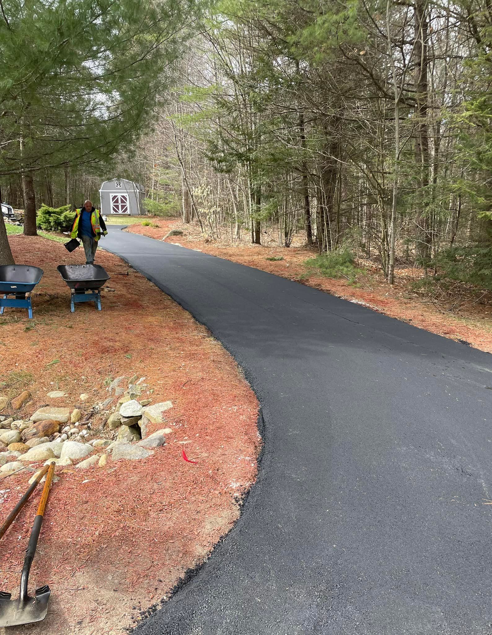 A worker in a high-visibility vest stands on a newly paved asphalt driveway in a wooded area with wheelbarrows nearby.