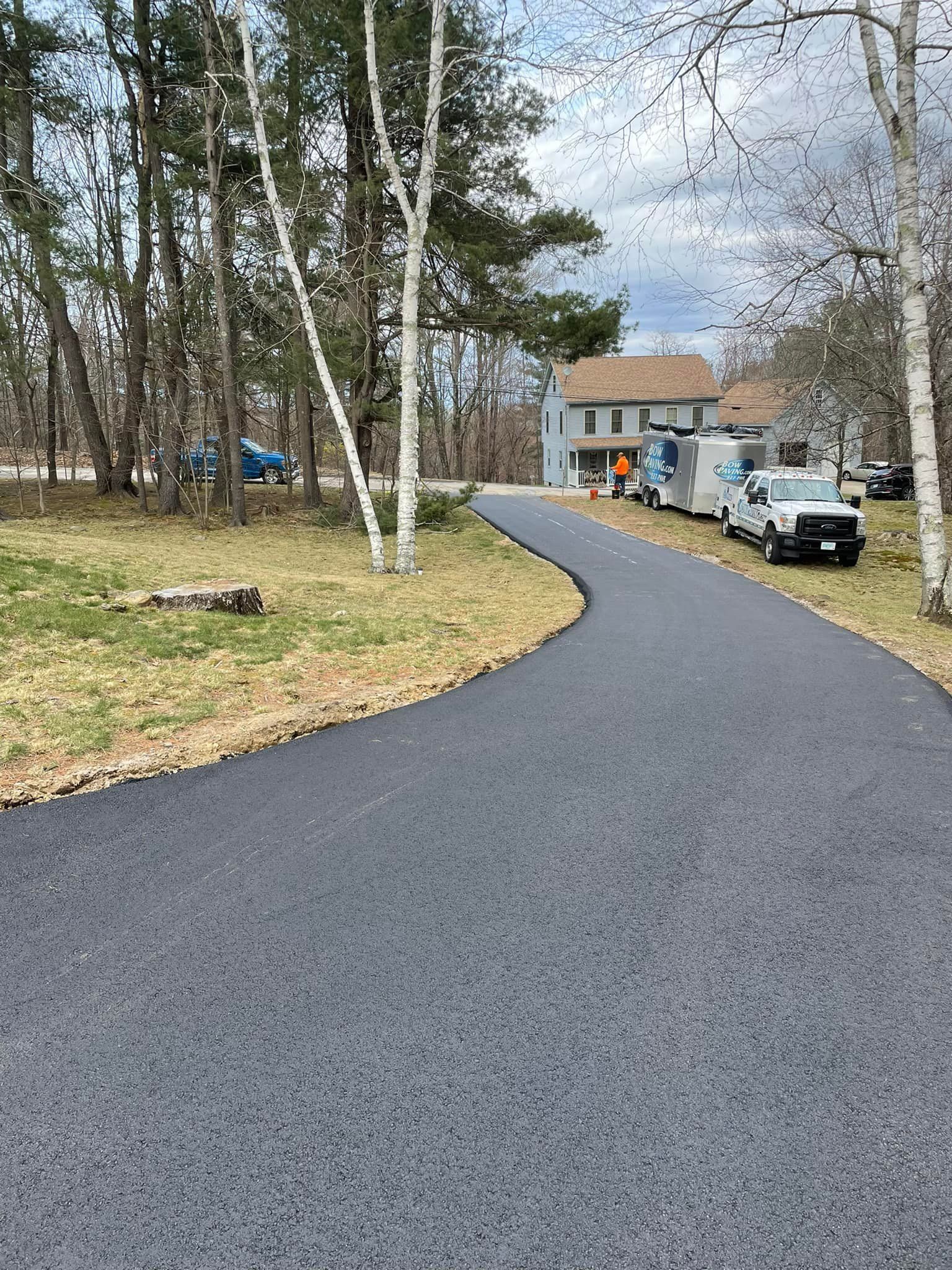 A freshly paved asphalt driveway winds through a grassy, wooded yard toward a two-story house with a work truck parked near.