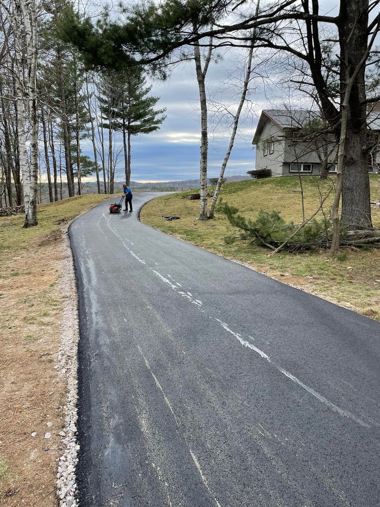 A person on a bicycle travels along a freshly paved, winding road bordered by trees and a house on a grassy hill.