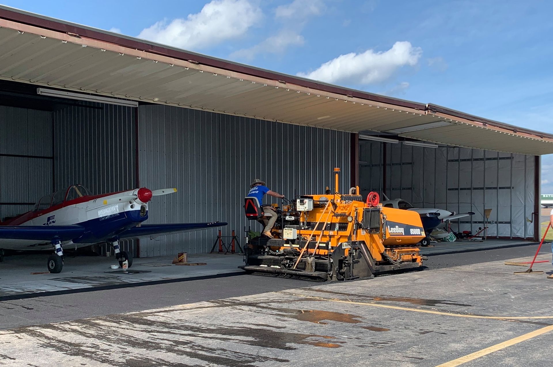 A red, white, and blue airplane parked in a hangar next to a yellow construction machine on a sunny day.