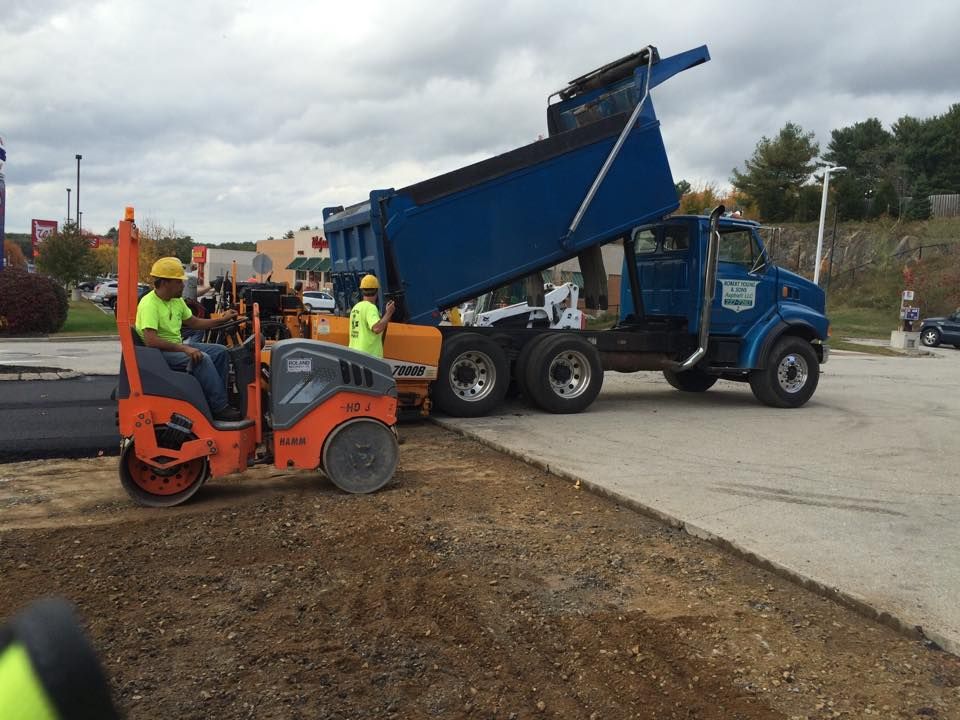 Construction workers operate an orange roller and a yellow paving machine next to a blue dump truck on an asphalt lot.