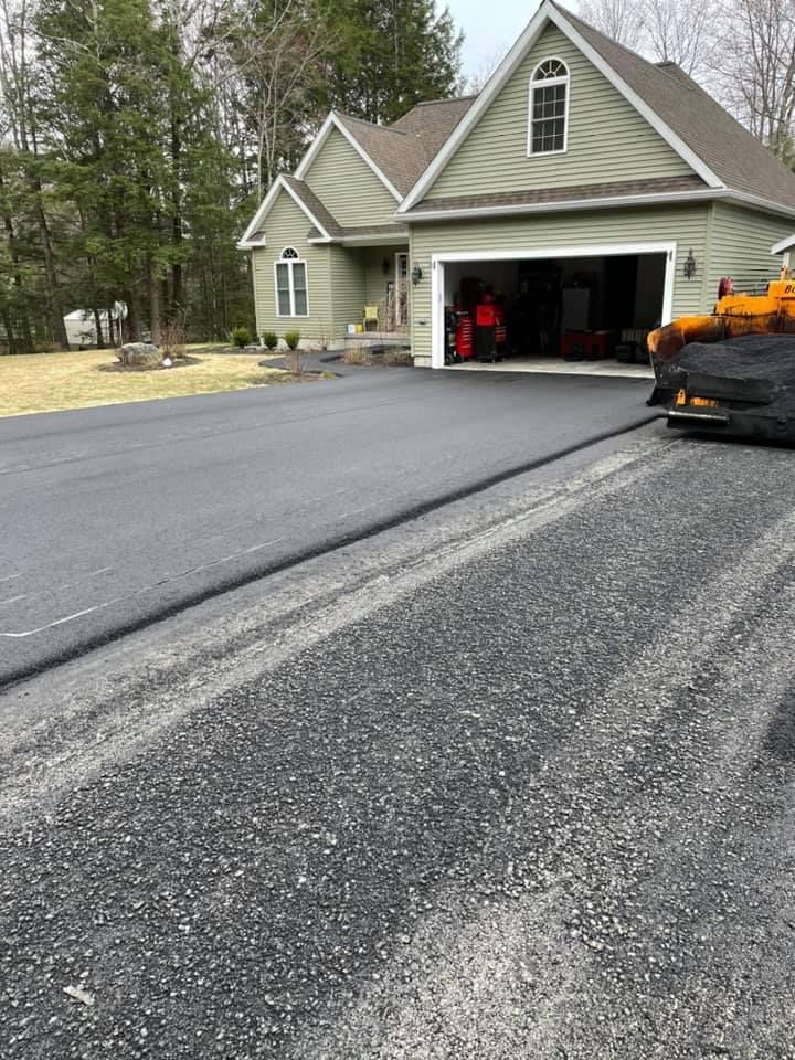 A paved driveway section newly installed next to a section of gravel, in front of a light green house with a garage.