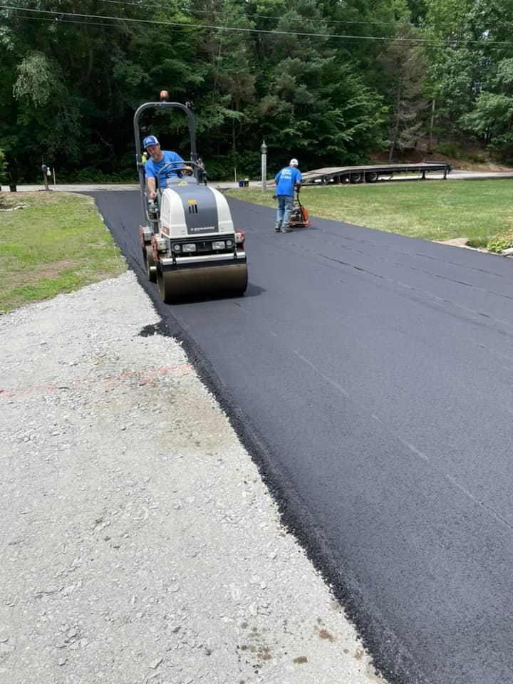 Two workers use construction machinery to pave a long, dark asphalt driveway in a grassy, wooded outdoor setting.