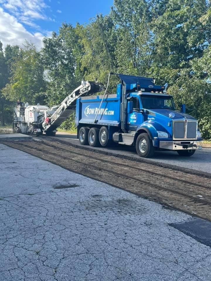A blue dump truck and a milling machine removing old asphalt from a road surrounded by trees under a blue sky.