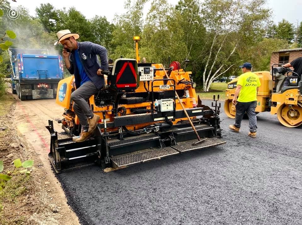 A person in a straw hat talks on a phone while operating an orange asphalt paver on a road construction site.