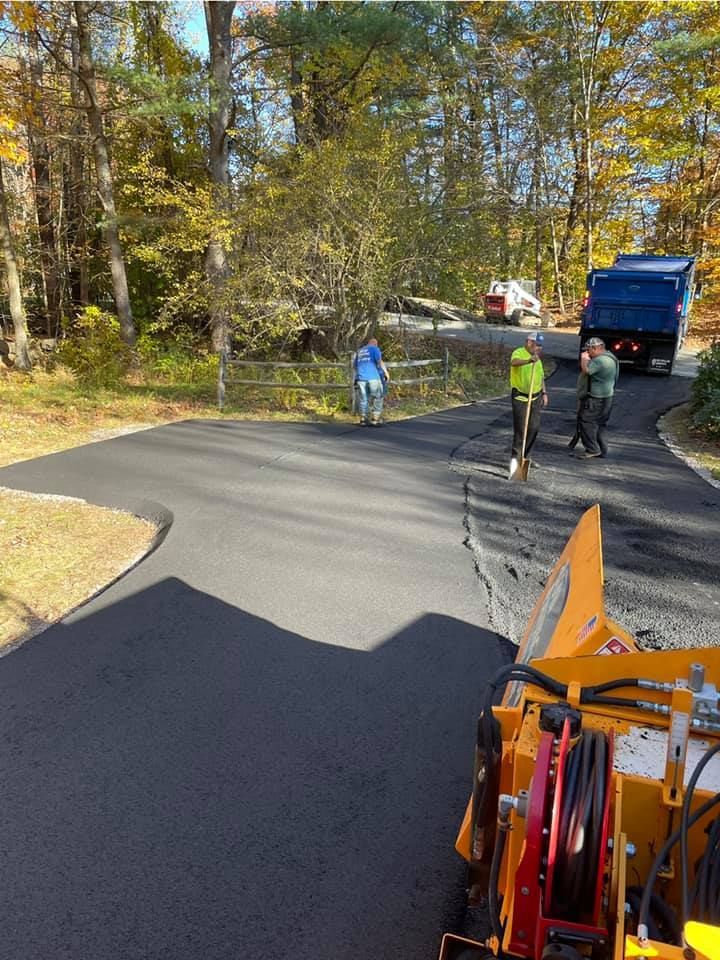 Three workers finish laying fresh black asphalt on a driveway surrounded by trees, with a dump truck parked nearby.