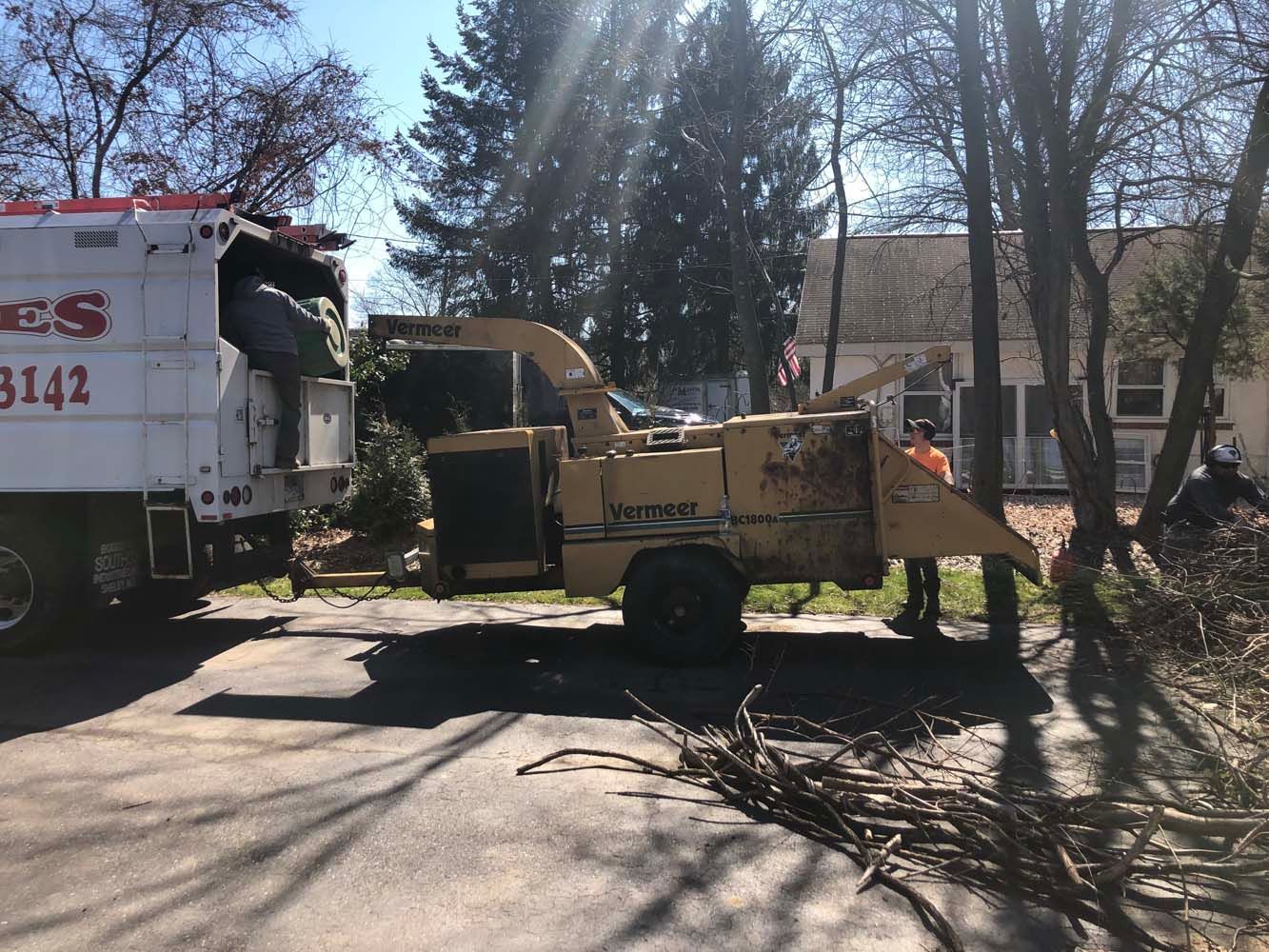 A tan wood chipper is attached to a white truck on a paved driveway, with a worker standing nearby.