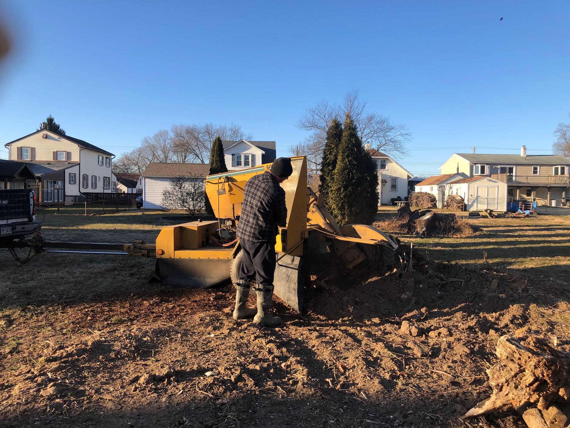 A person in a dark jacket uses a yellow stump grinder in a yard with houses in the background on a sunny day.