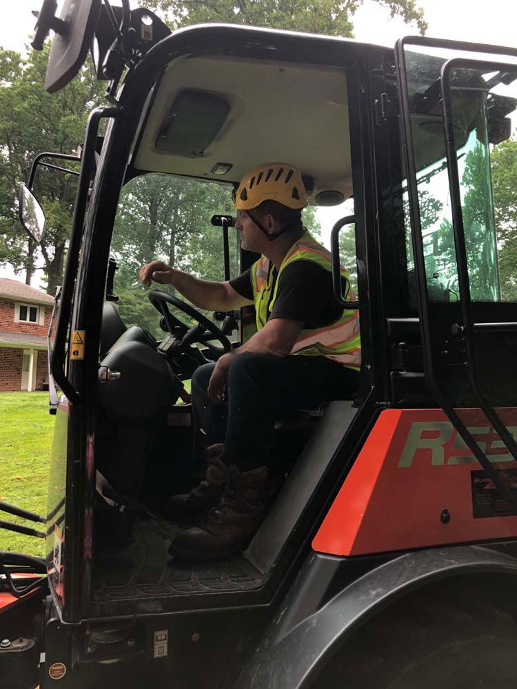 A construction worker in a high-visibility vest and yellow hard hat sits in the operator cab of heavy machinery.