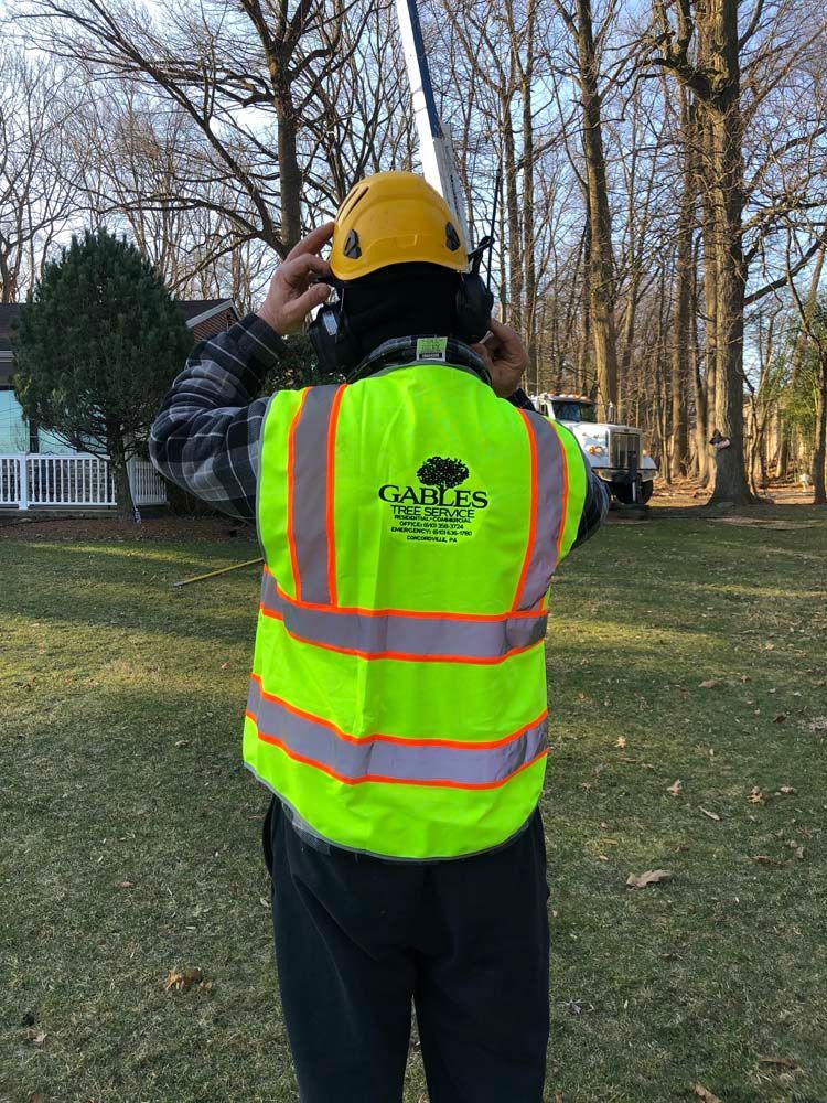 A worker in a high-visibility vest and yellow hard hat stands outdoors, facing a boom lift in a wooded residential yard.