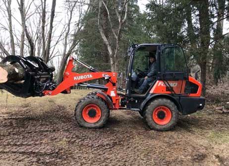 A bright orange Kubota wheel loader equipped with a grapple attachment carries a large log through a wooded area.