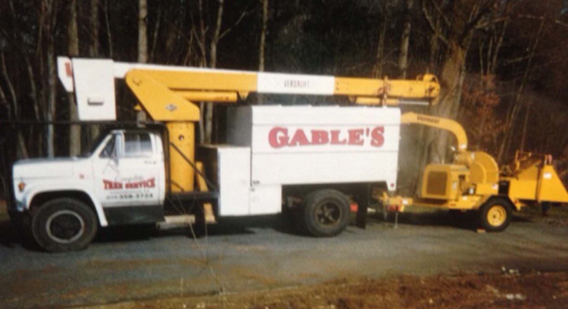 White bucket truck and yellow wood chipper parked against a dark, wooded background. The truck is labeled 