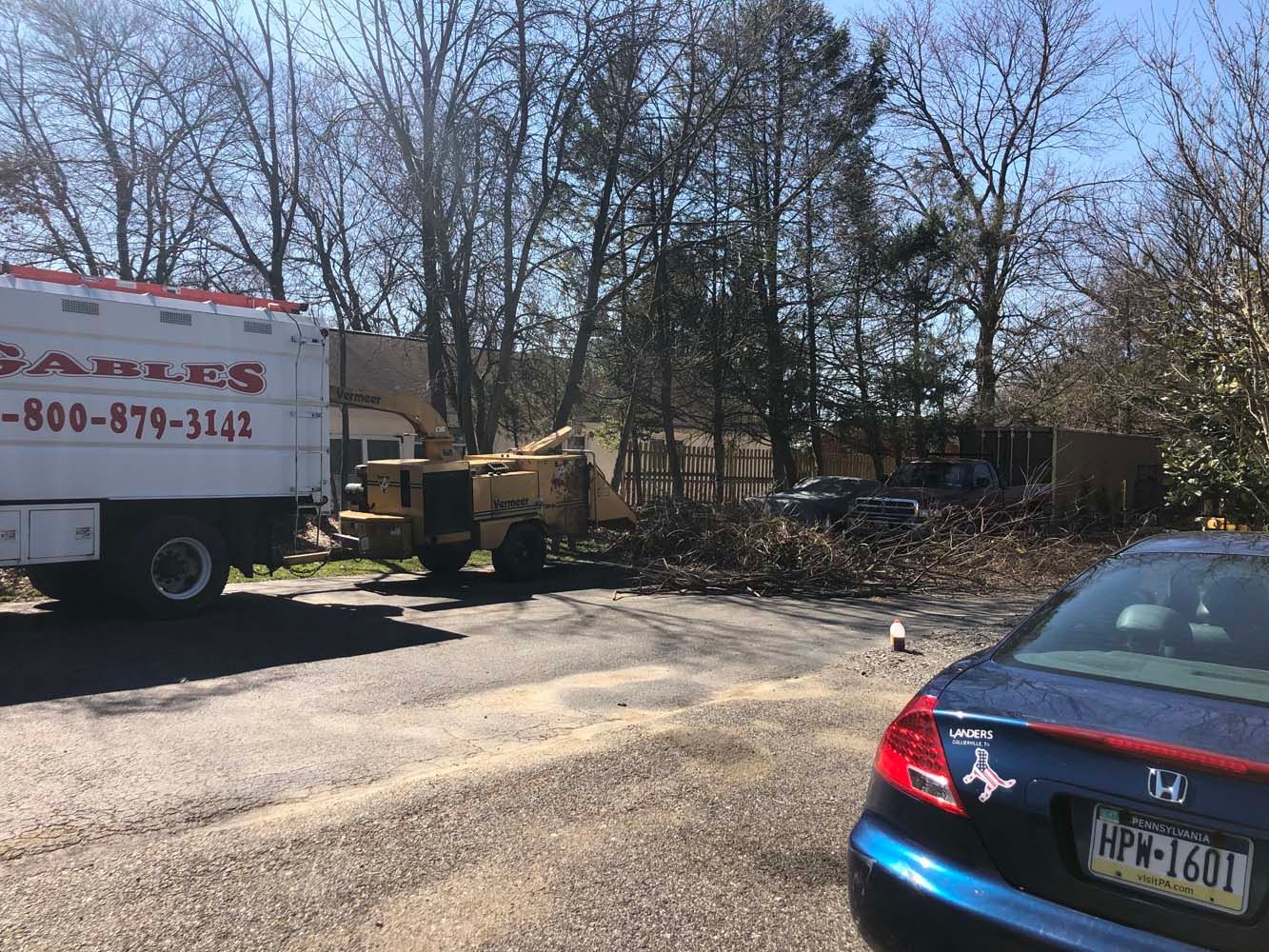 A wood chipper attached to a truck, parked on a gravel lot near a blue car, with trees in the background.