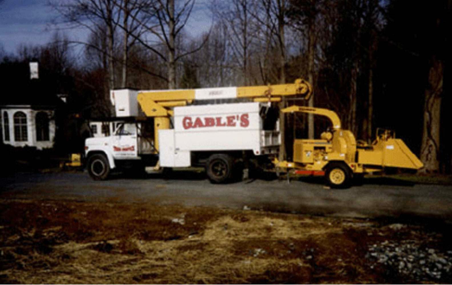 A yellow and white Gable’s tree service truck with an attached wood chipper parked in front of a house.