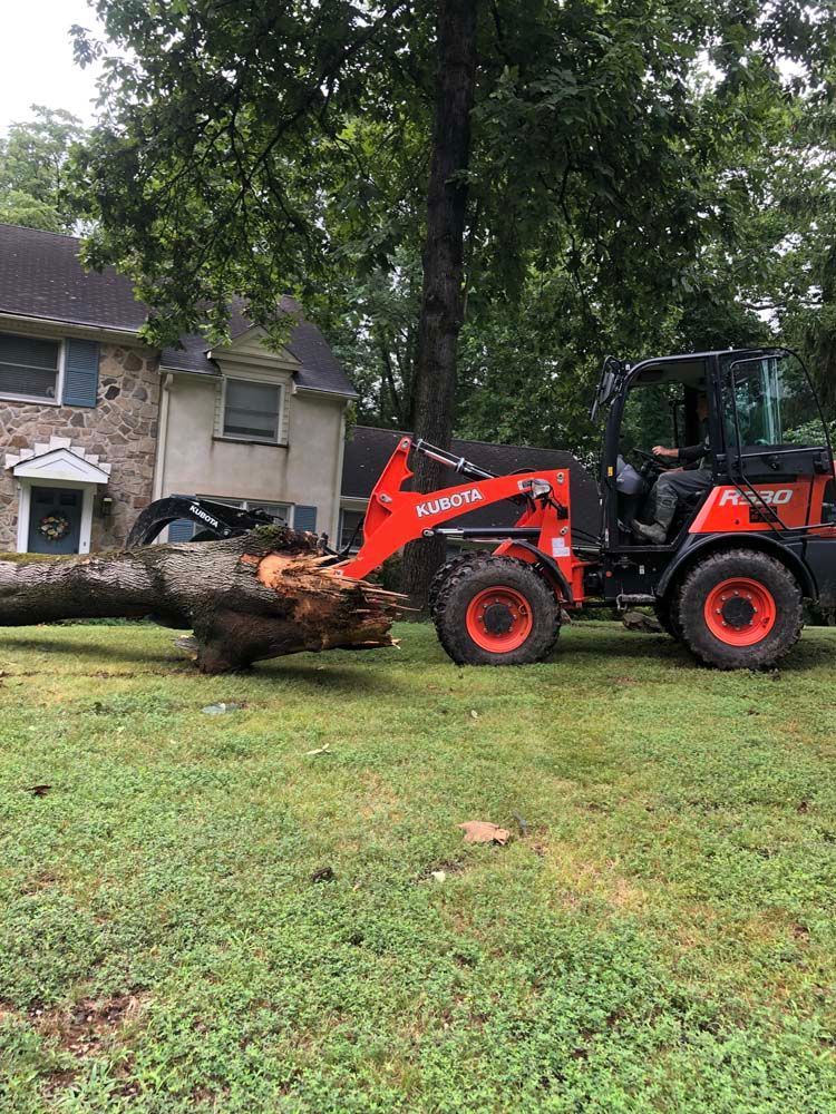 An orange Kubota wheel loader moves a large, cut tree trunk across a grassy lawn in front of a multi-story stone house.