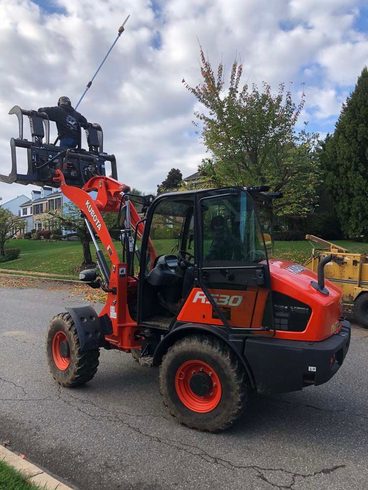 A person stands on the elevated grapple attachment of an orange Kubota wheel loader parked on an asphalt street.