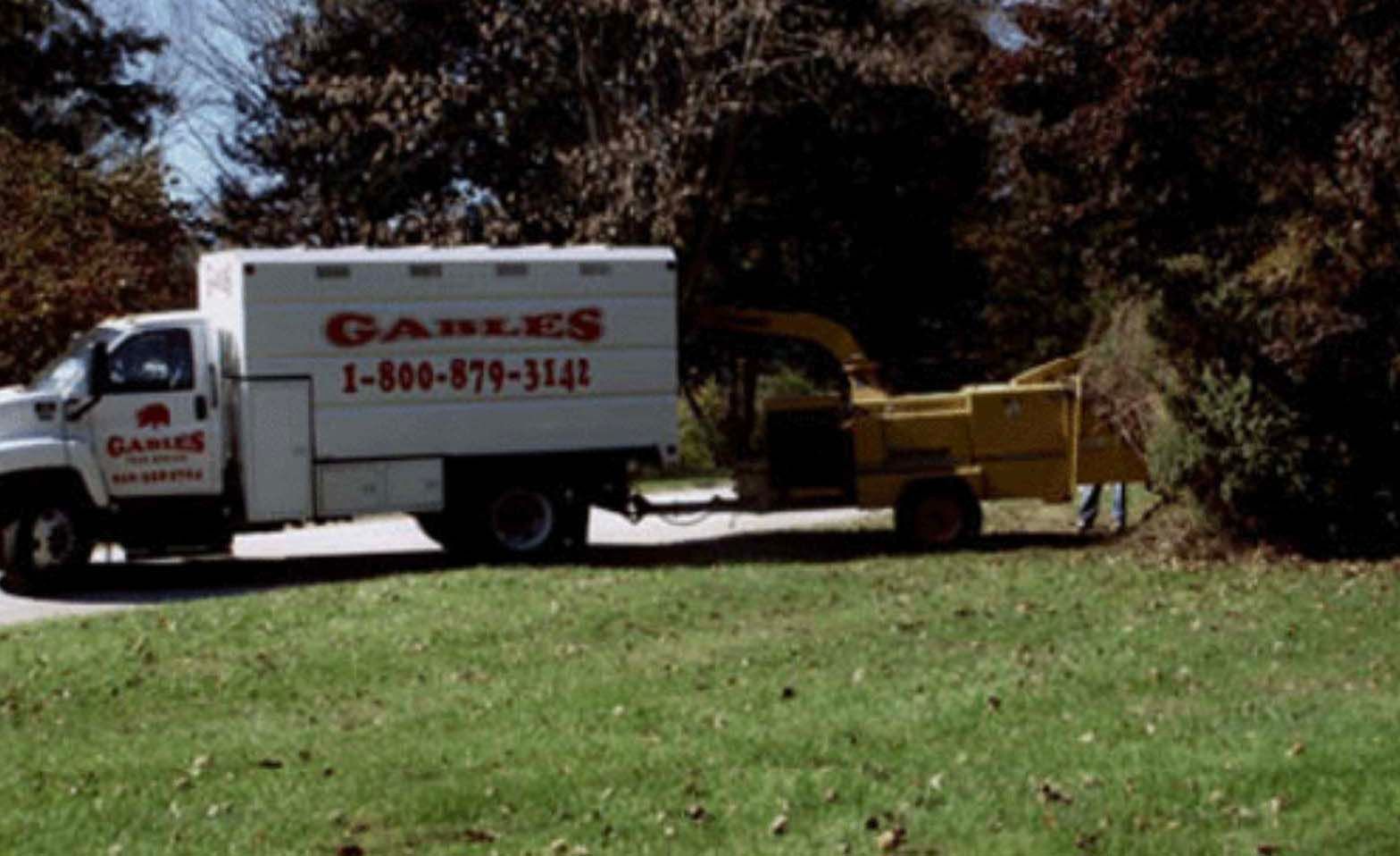 A white Gables service truck with a wood chipper attached, parked on a grassy area next to trees.