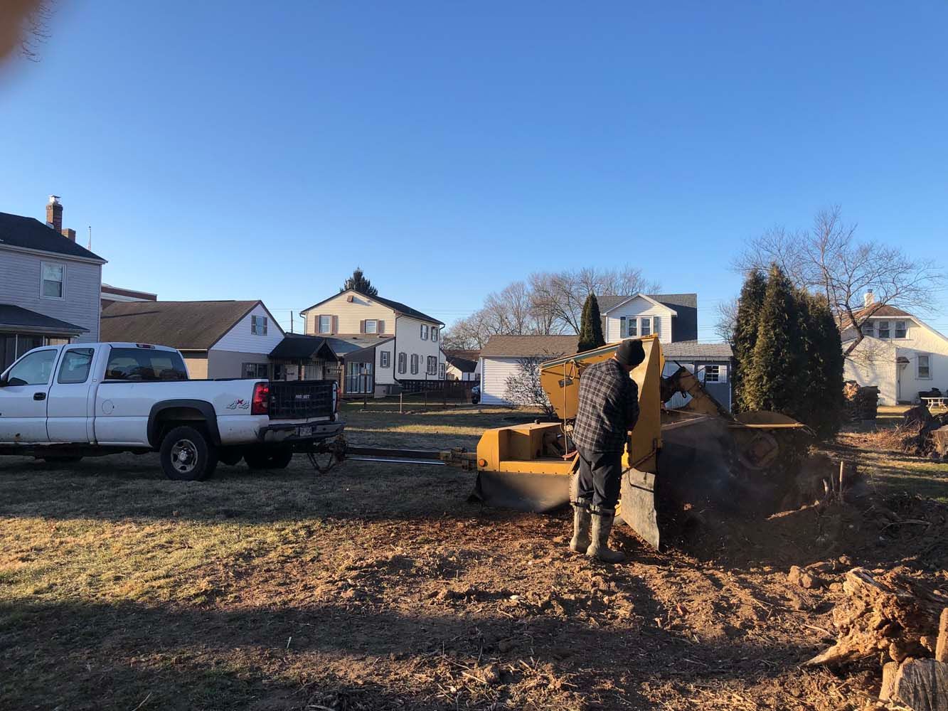 A person operates a yellow piece of construction machinery to clear land in a residential neighborhood near a white truck.