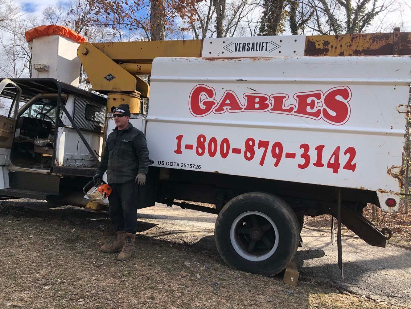 A person wearing a jacket and cap holds a chainsaw next to a white bucket truck labeled 