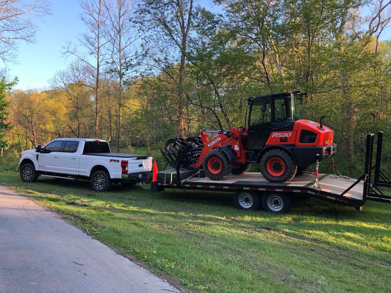 A white pickup truck parked on grass pulling a flatbed trailer loaded with a bright orange Kubota wheel loader.