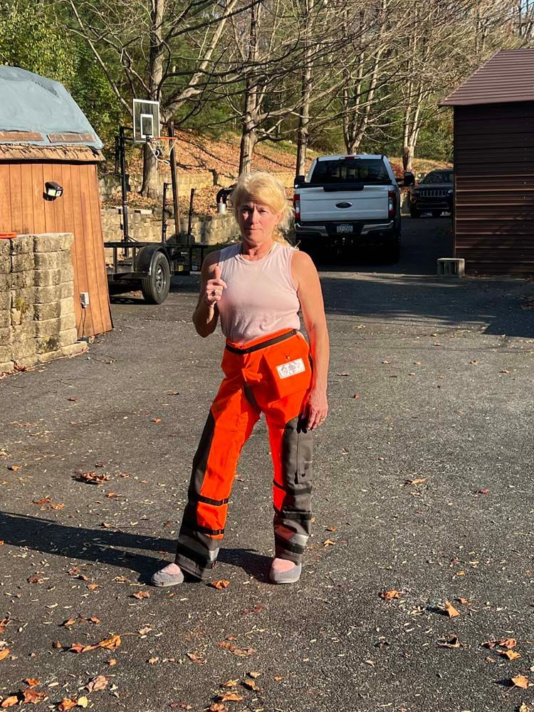 A person in bright orange protective work pants and a light tank top stands outdoors giving a thumbs up.