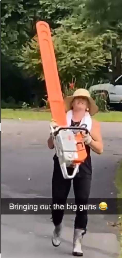 A person wearing a straw hat and boots walks outdoors while holding an extremely large orange chainsaw.