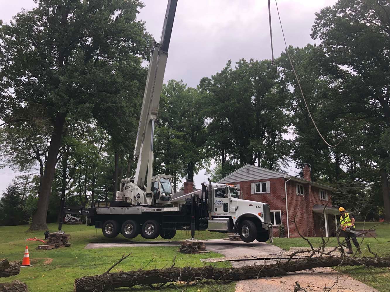 A crane truck parked on a residential driveway, lifting a cut tree branch near a brick house while a worker stands nearby.