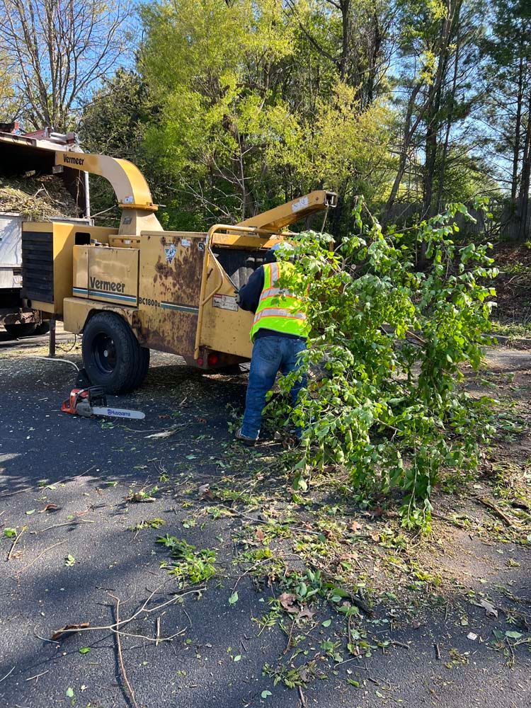 A worker in a high-visibility vest feeds branches into a large yellow industrial wood chipper outdoors.