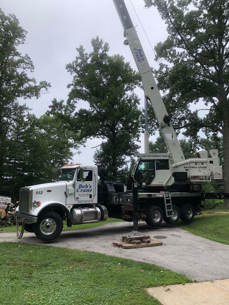 A white boom truck crane parked on a paved driveway surrounded by trees.