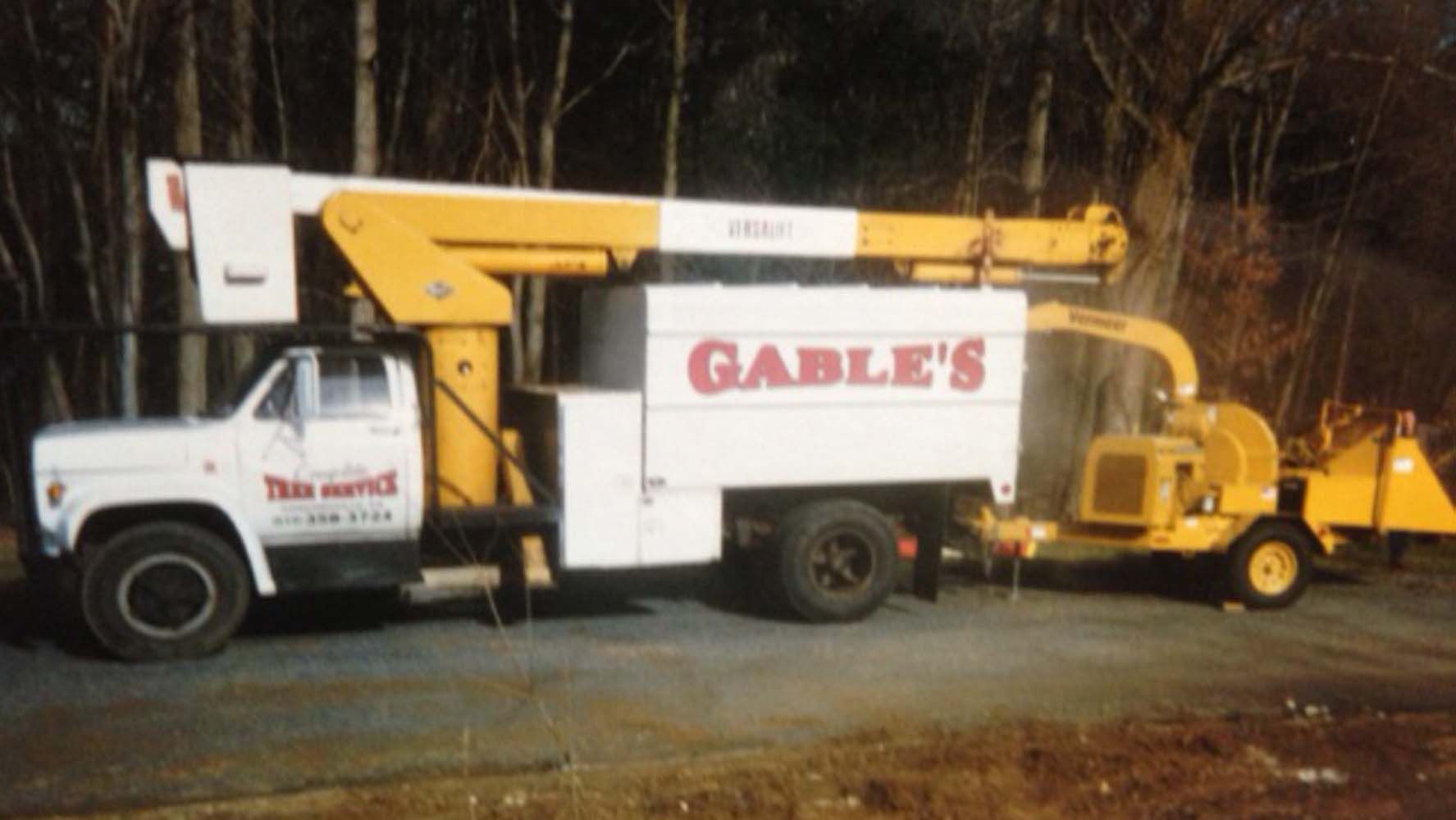 A white and yellow Gable’s tree service bucket truck towing a wood chipper parked in front of trees.