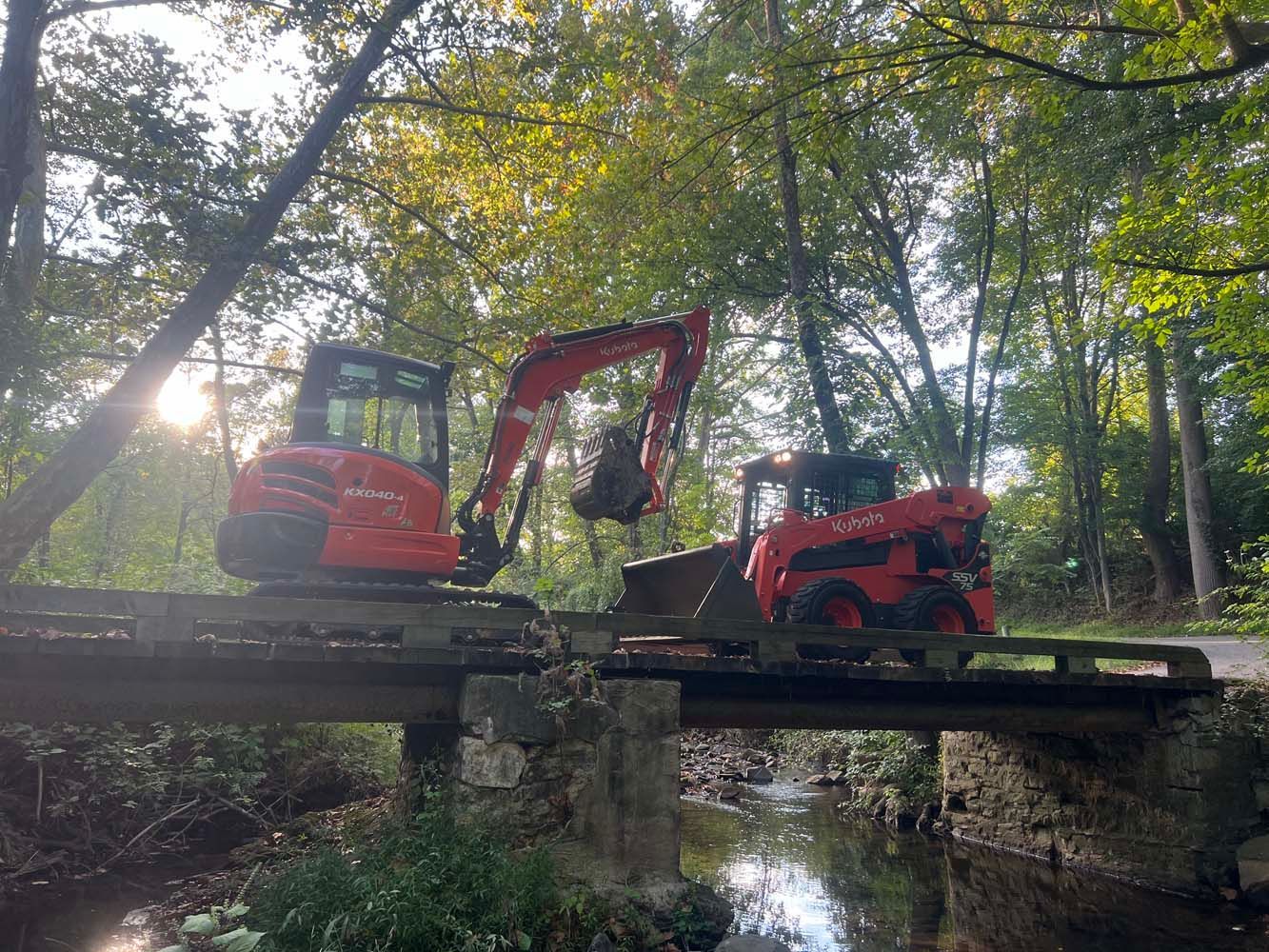 Red mini-excavator and skid steer parked on a stone bridge over a forest stream at sunset.