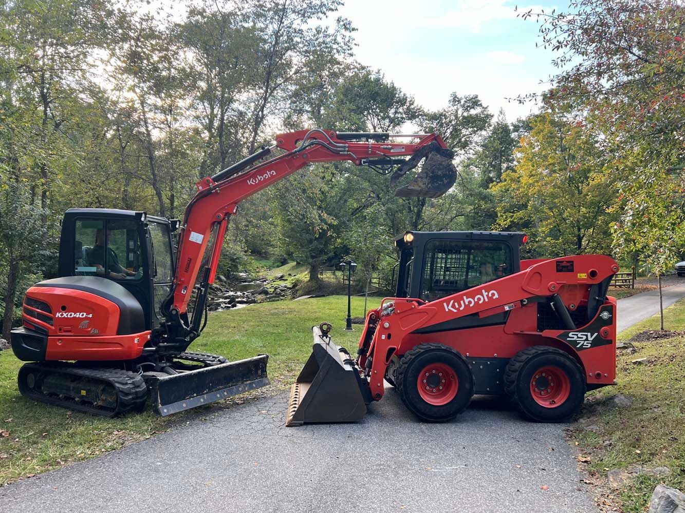 A red Kubota excavator and a red Kubota skid steer loader parked side-by-side on an outdoor paved path.
