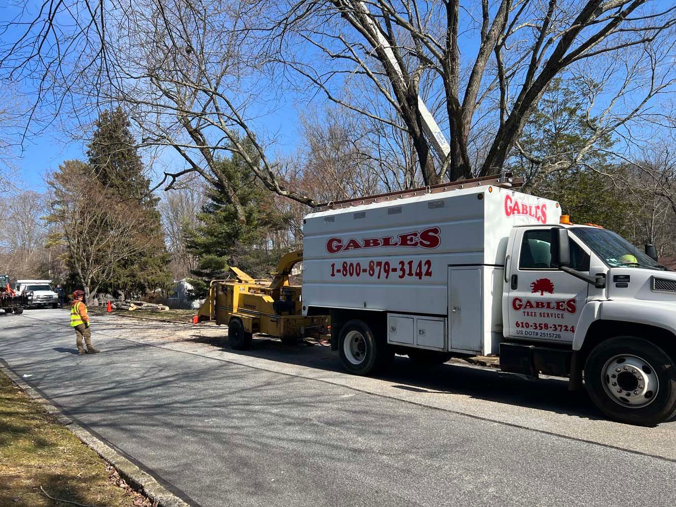 A white Gables utility truck parked on a road next to a yellow wood chipper, with a worker standing nearby.