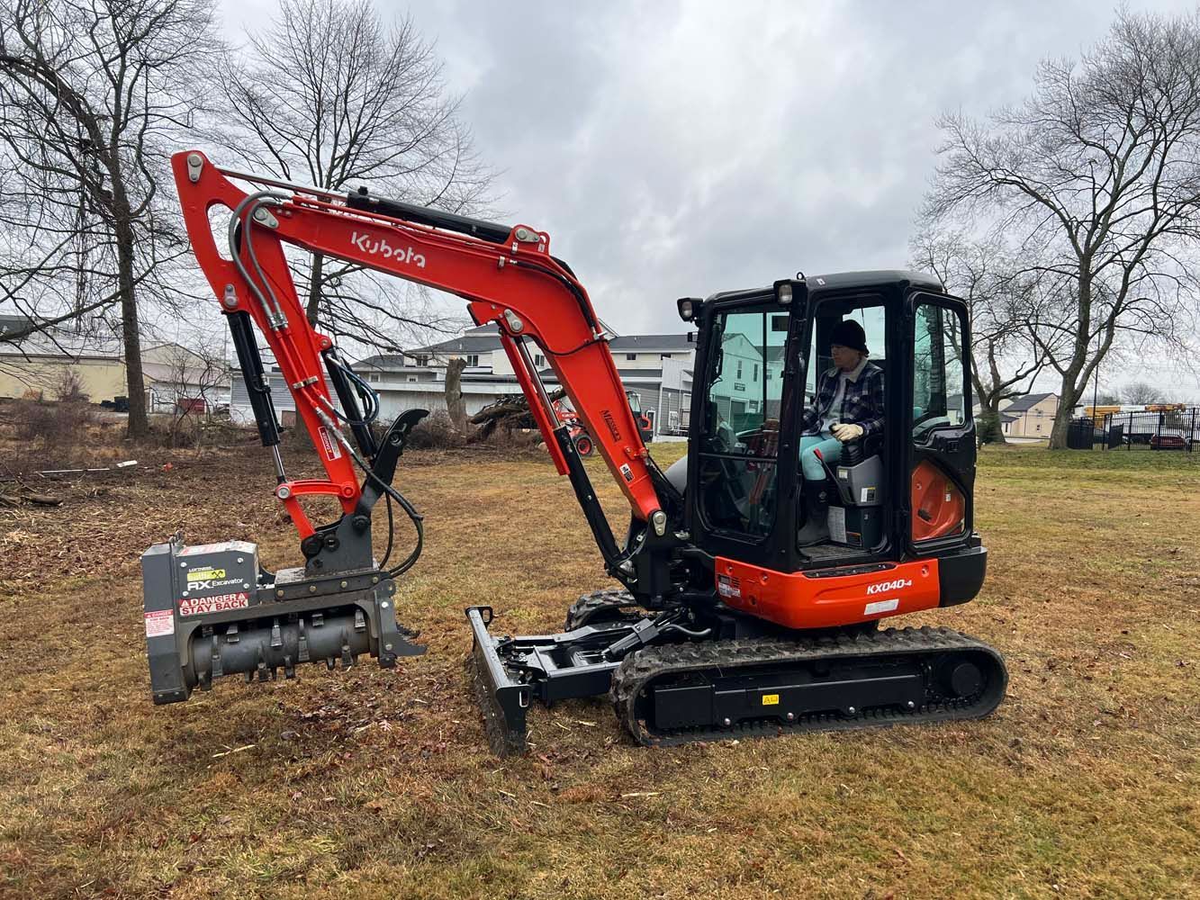 A red Kubota compact excavator with a mulching attachment operates in an open, grassy field under a cloudy sky.