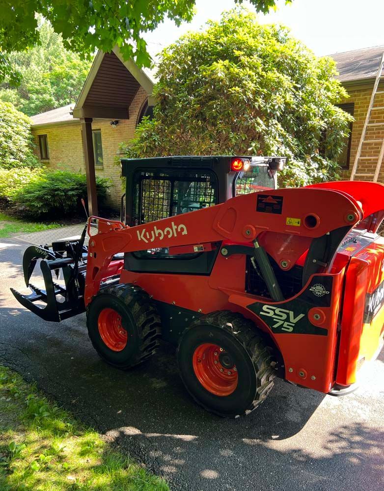 A red Kubota SSV75 skid steer loader with a grapple attachment parked on a paved driveway in front of a house.