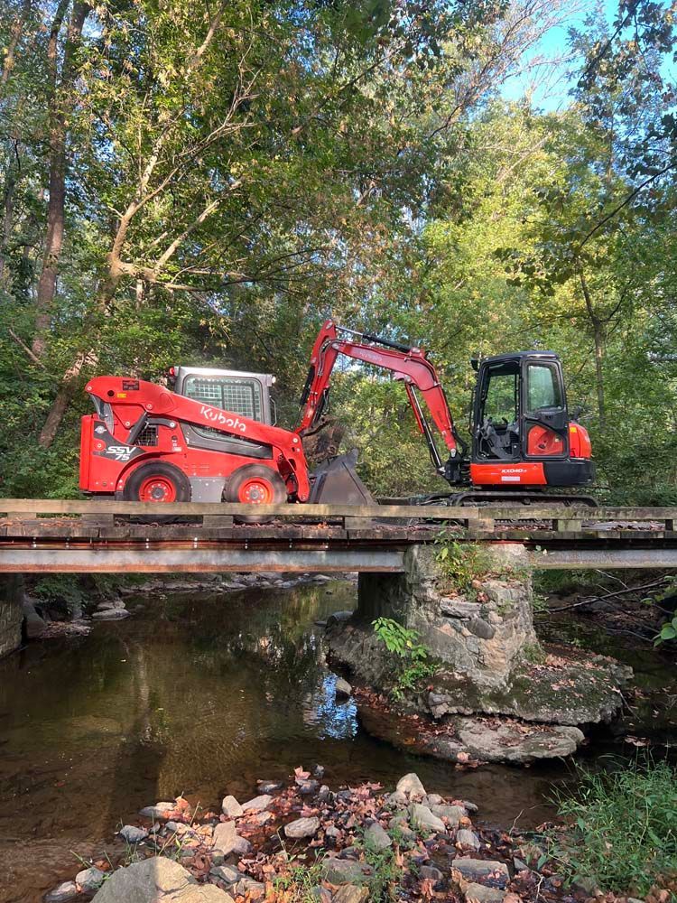 A red skid steer and an excavator parked on a weathered wooden bridge over a small creek in a wooded area.