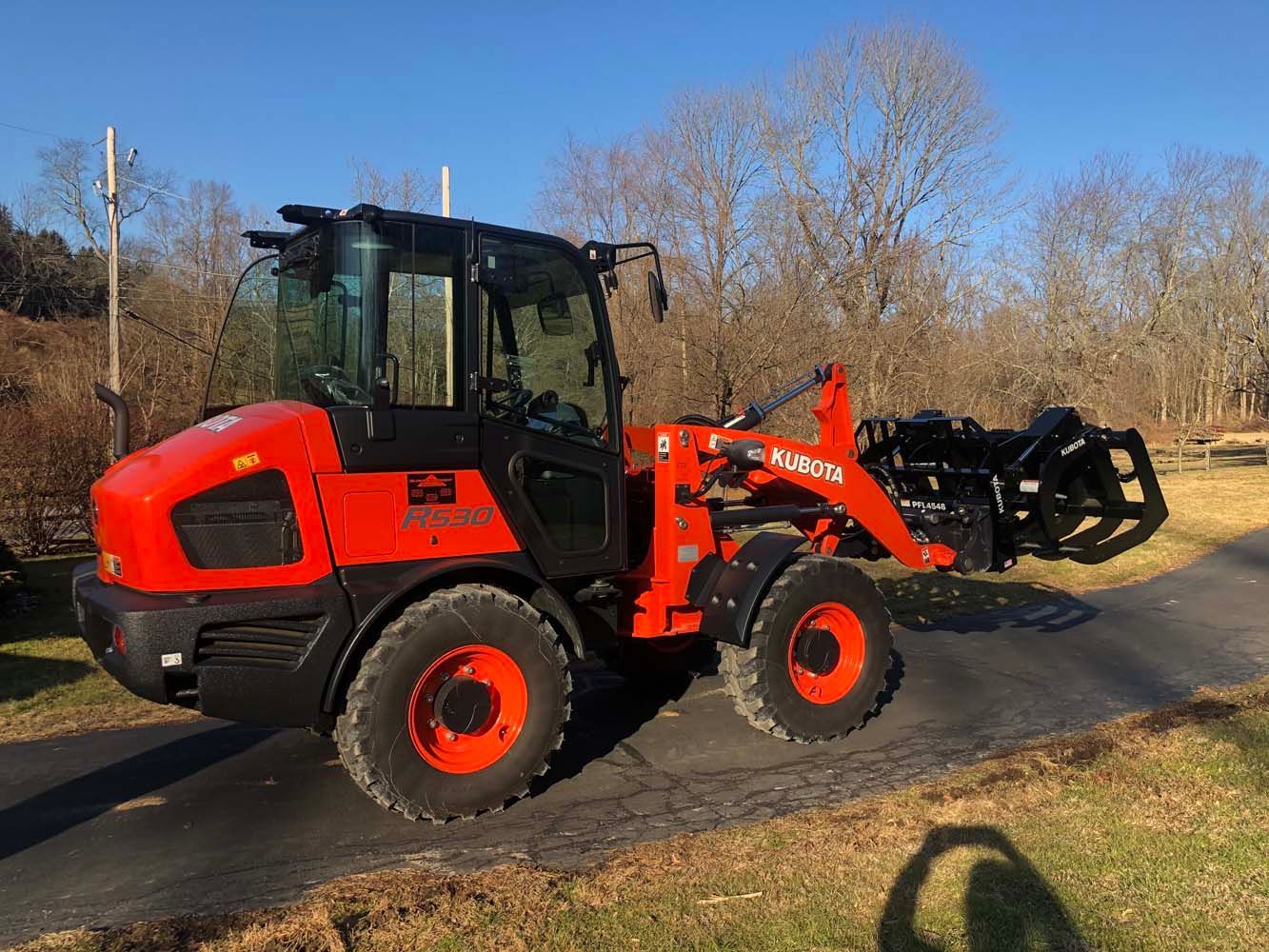 An orange and black Kubota wheel loader equipped with a grapple attachment, parked on an asphalt path in a grassy area.