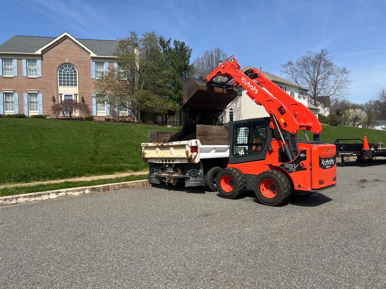 An orange Kubota skid-steer loader dumps dirt into the back of a white dump truck in front of a house on a sunny day.
