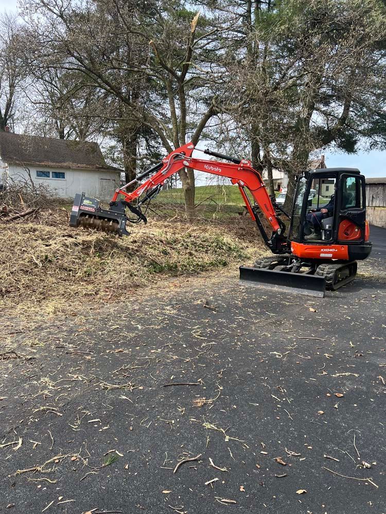 An orange Kubota excavator with a forestry mulcher attachment working on a pile of brush near trees and a building.