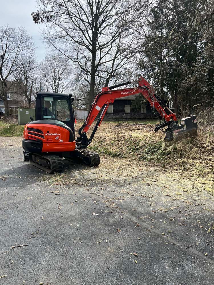 An orange Kubota excavator parked on a paved lot, clearing brush and debris near a wooded area.