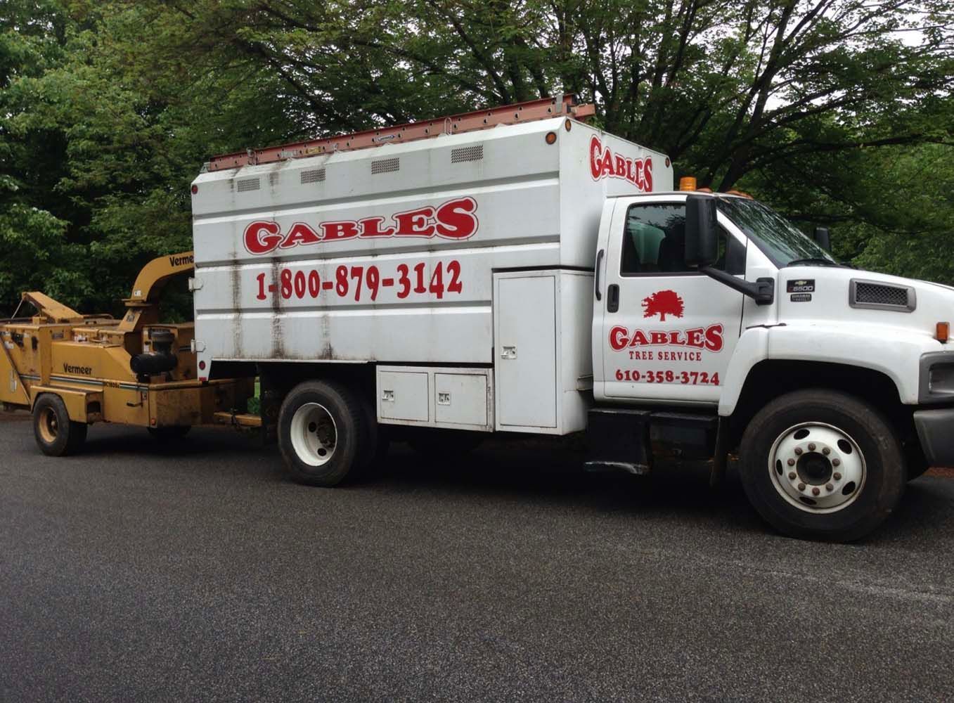 A white Gables tree service truck with an attached yellow wood chipper parked on an asphalt road near green trees.